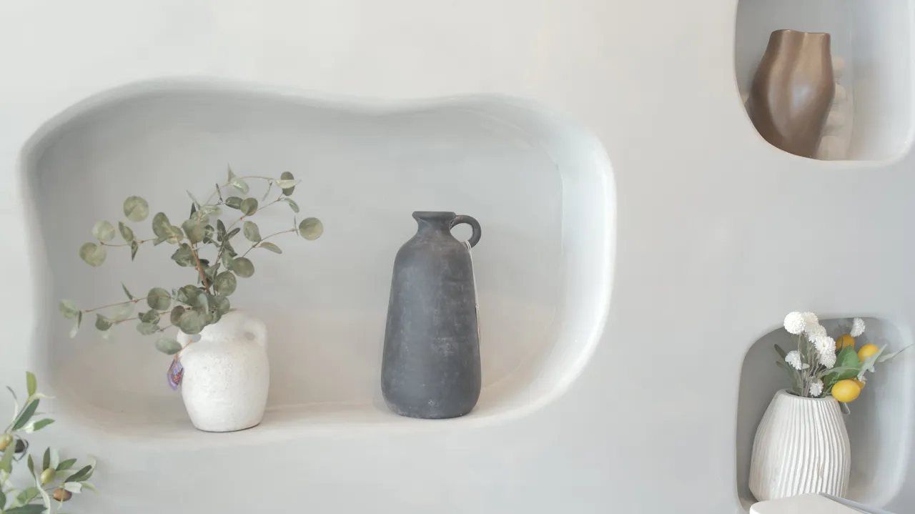 A black ceramic jug and a white vase of eucalyptus in a smooth rounded alcove carved into a white plaster wall.